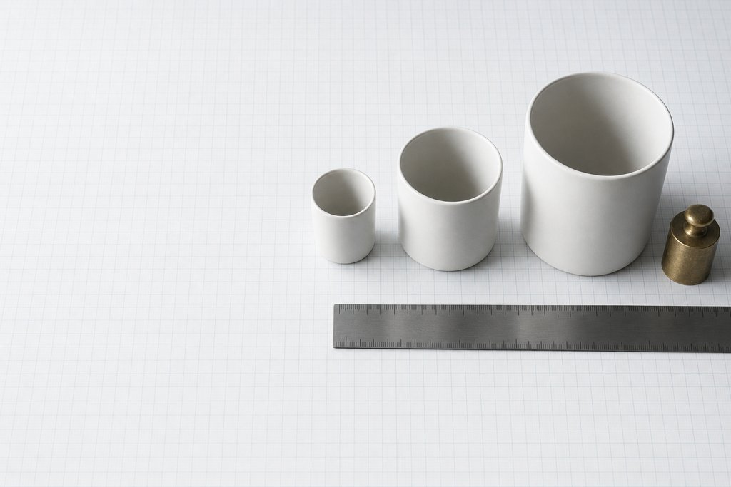 Three plain ceramic cups of increasing size on graph paper with a brass balance weight and a steel rule